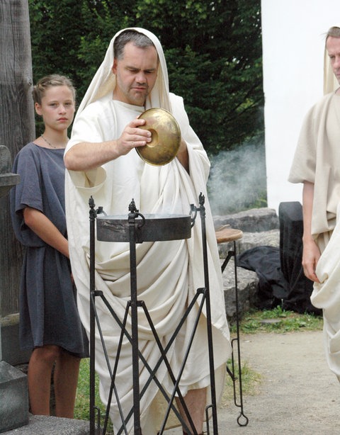 Spielszene: Ein Mann in eine Toga gehüllt, schüttet etwas in ein Feuer.