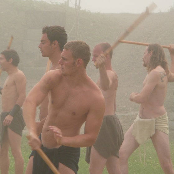 Mehrere spärlich bekleidete junge Männer bei Kampfübungen mit Stöcken.