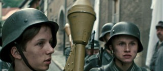 Mehrere Jungen in Uniformen laufen durch eine Straße. Einer von ihnen trägt eine Panzerfaust.