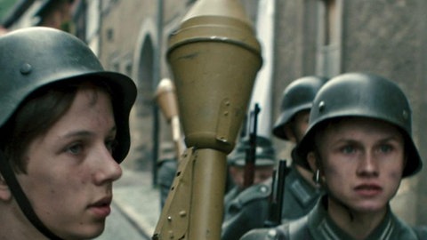 Mehrere Jungen in Uniformen laufen durch eine Straße. Einer von ihnen trägt eine Panzerfaust.