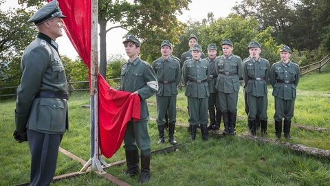 Soldat vor Jungen in Uniform. Dazwischen eine rote Fahne.