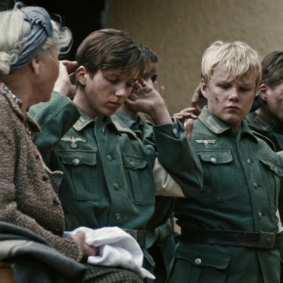 Jungen in Uniform haben die Hände erhoben, daneben eine Frau mit weißem Tuch.