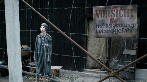 Junges Mädchen in Häftlingskleidung hinter Drahtzaun schaut traurig nach oben.