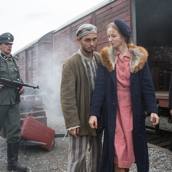 Gut gekleidete junge Frau am Zug, hinter ihr ein Mann in Sträflingskleidung und ein SS-Mann mit Waffe.