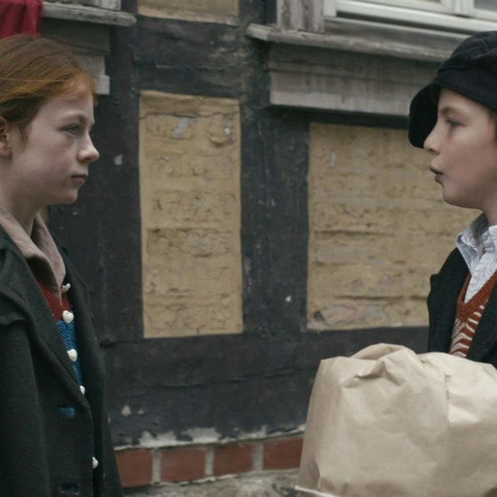 Greta und Anton sprechen mit ernsten Mienen miteinander, im Hintergrund zu sehen: eine Hakenkreuzfahne.