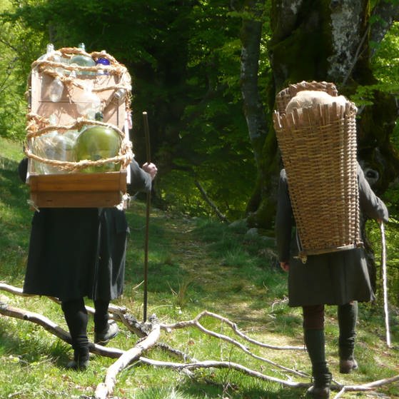 Glasträger mit Holzgestell und Glaswaren auf dem Rücken mit Begleiter, der einen Korb auf dem Rücken trägt – Ansicht von hinten (Foto: Tilmann Büttner)