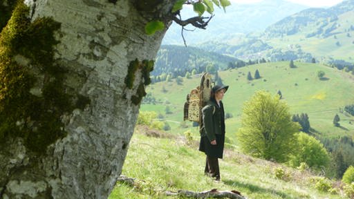 Mann in Hügellandschaft, trägt Gestell mit Uhren auf dem Rücken