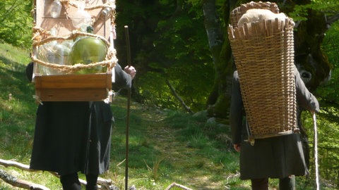 Glasträger mit Holzgestell und Glaswaren auf dem Rücken mit Begleiter, der einen Korb auf dem Rücken trägt – Ansicht von hinten