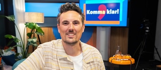 Host Gero sitzt vor einem Bildschirm mit der Aufschrift "Komma klar!" und lacht in die Kamera