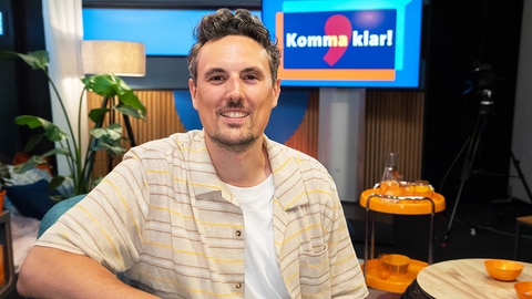 Host Gero sitzt vor einem Bildschirm mit der Aufschrift 