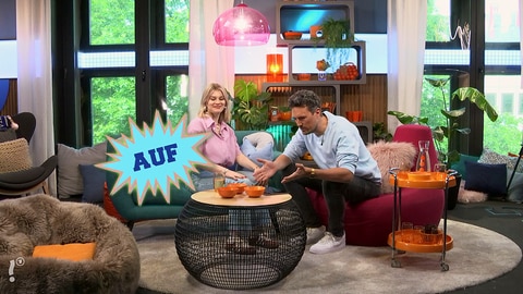 Host Gero sitzt mit einer Frau im Studio an einem Tisch, eingeblendet das Wort 