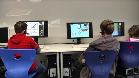 Drei Kinder sitzen mit dem Rücken zur Kamera vor Computern und spielen das Lernspiel Deutsch lernen mit Mumbro und Zinell.