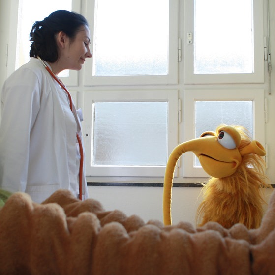 Eine Ärztin im weißen Kittel unterhält sich mit Zinell; Mumbro liegt auf einem Bett.