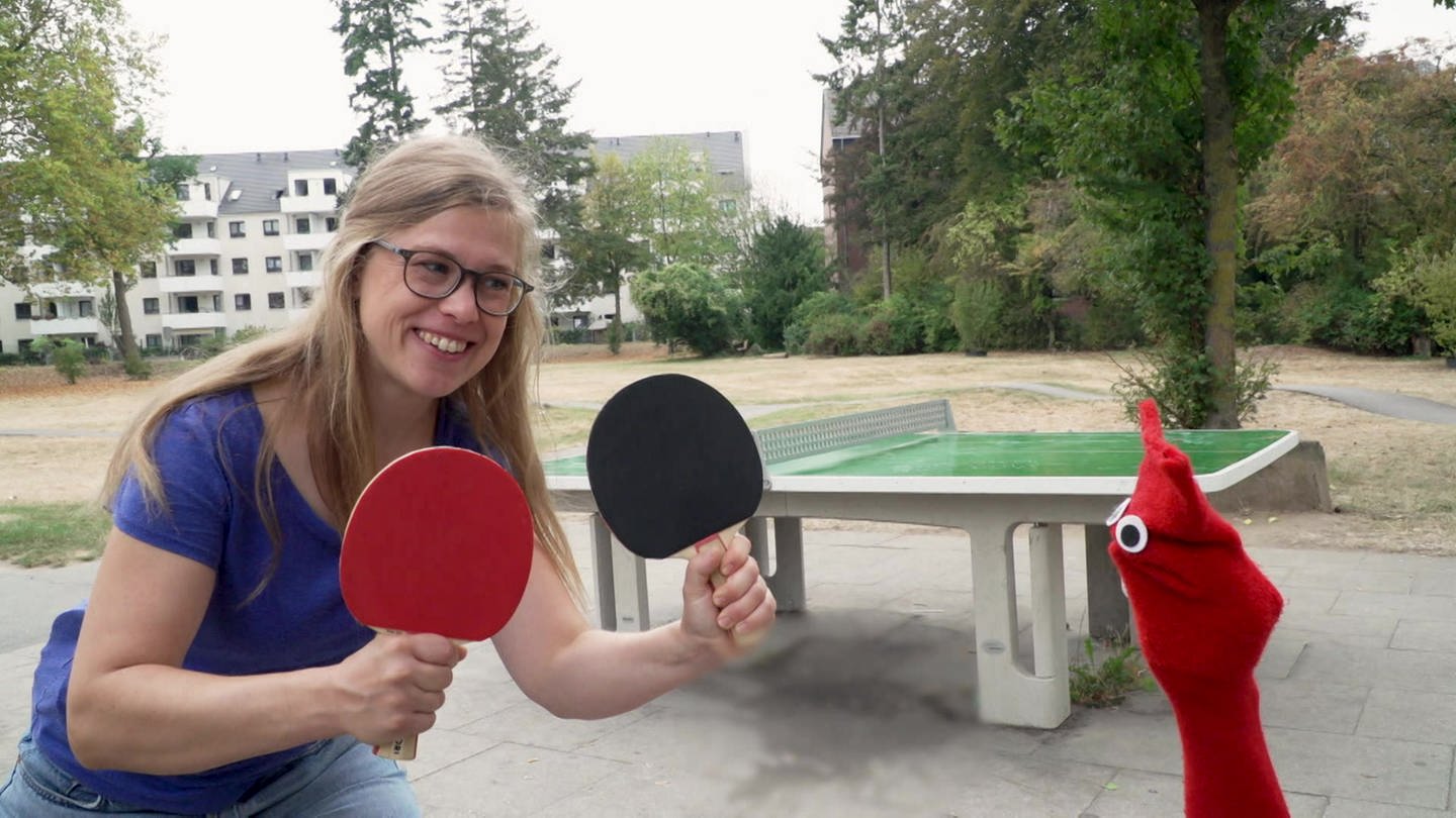 Esther und die rote Socke stehen vor einer Tischtennisplatte. Esther hält zwei Schläger in der Hand. (Foto: WDR)