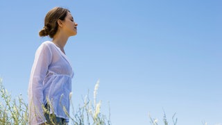 Eine Frau atmet tief auf einer Wiese vor bauem Himmel: Ohne Sauerstoff können wir nicht leben.