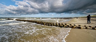 Wellen brechen sich an Buhnen an einem Strand. Die Reihen aus Holz ragen ins Meer, bremsen das Abfließen des Wassers und unterstützen dadurch die Ablagerung von Schlick. 