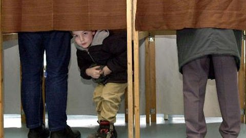 zwei Wahlkabinen in den mit zugezogenen Vorhängen, unter einem der Vorhänge schaut ein kleiner Junge hervor (Foto: dpa)