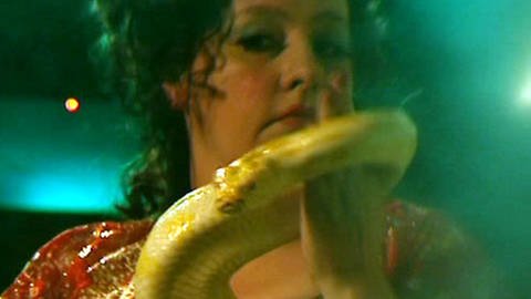 Ein exotisch wirkende Frau, von der Kopf und Schultern zu sehen sind, hält eine gelbe Schlange vor sich. (Foto: SWR – Print aus der Sendung)