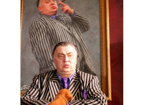An einem Schreibtisch sitzt ein etwas korpulenter Mann mit grau-gestreiftem Jackett sowie lila Hemd und Krawatte. Hinter ihm ein lebensgroßes Gemälde des Mannes in der gleichen Kleidung und mit einer dicken Zigarre in der Hand. (Foto: Daniel Engstler)