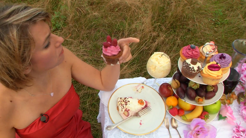 Eine Frau in rotem Kleid sitzt an einem Tisch mit Kuchen und Obst und hält einen Cupcake in der Hand. Essen im Überfluss hat Folgen für Mensch und Umwelt.
