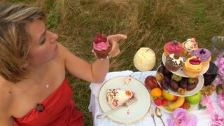 Eine Frau in rotem Kleid sitzt an einem Tisch mit Kuchen und Obst und hält einen Cupcake in der Hand. Essen im Überfluss hat Folgen für Mensch und Umwelt.