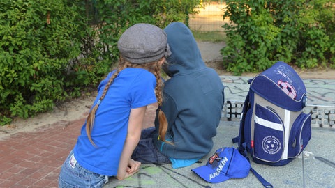 Zwei Schulkinder sitzen mit dem Rücken zum Betrachter auf einer Tischtennisplatte, neben ihnen ein Schulranzen