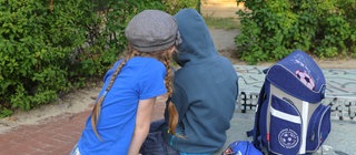 Zwei Schulkinder sitzen mit dem Rücken zum Betrachter auf einer Tischtennisplatte, neben ihnen ein Schulranzen