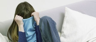 Mädchen sitzt auf Sofa und verdeckt ihr Gesicht mit ihrem Pulli.