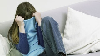 Mädchen sitzt auf Sofa und verdeckt ihr Gesicht mit ihrem Pulli.