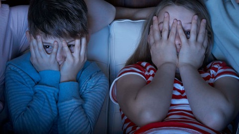 Zwei Kinder sitzen im Schein eines Fernsehers und gucken durch die Hände, mit denen sie sich die Augen halb zuhalten
