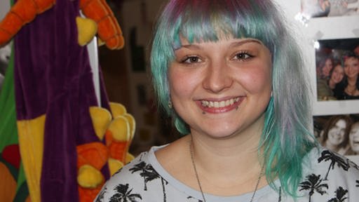 Eine junge Frau mit blau-lilalen Haaren und Piercings lächelt in die Kamera
