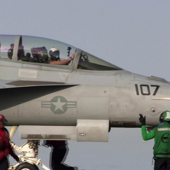 Mehrere Männer stehen neben einem Militär Jet. Zwei Piloten winken aus dem Jet.