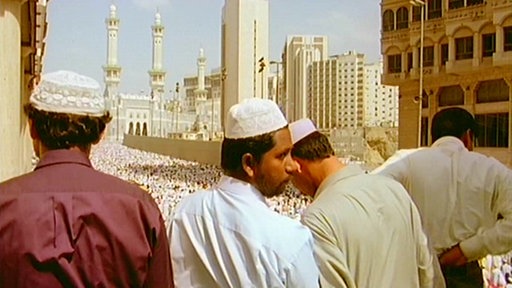 Mehrere Männer mit weißen Kopfbedeckungen stehen auf einem Balkon und schauen über eine Menschenmenge.