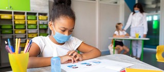 Ein Mädchen mit Mundschutz am Tisch in einem Klassenzimmer. Im Hintergrund ein anderes Kind und eine Frau. Distanz und Isolation in der Pandemie.