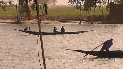 Silberner Fluss, darauf als Silhouette langes schmales Boot mit fünf Menschen. Im Vordergrund Sand, im Hintergrund grüne Felder.