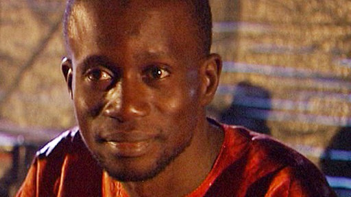 Koraspieler im hellroten Hemd sitzt vor seiner Kora im dunklen Musikclub