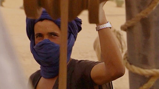 Kamele stehen dicht gedrängt in der Sandwüste an zwei Bäumen. Links von ihnen: ein Brunnen