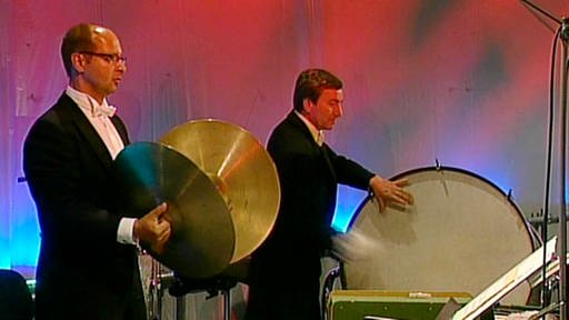 Zwei dunkel gekleidete Musiker spielen Becken und große Trommel. Der Hintergrund ist blau und rot beleuchtet. (Foto: SWR)