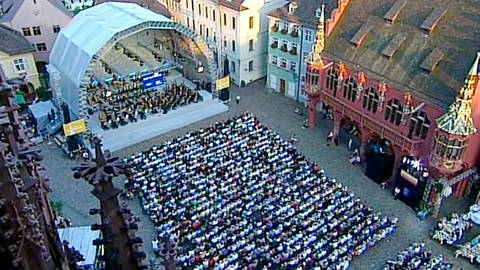 Vom Freiburger Münster aus gesehen: die überdachte Bühne mit dem Orchester, davor in Stuhlreihen das Publikum. Rechts von der Bühne sind historische Bauten zu erkennen, links angeschnitten das Freiburger Münster. (Foto: SWR)