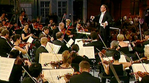 Dirigent Sylvain Cambreling steht vor dem Sinfonieorchester und dirigiert. Im Vordergrund die Streicher. Im Hintergrund ist durch die Abenddämmerung Teil einer Hausfassade zu sehen.