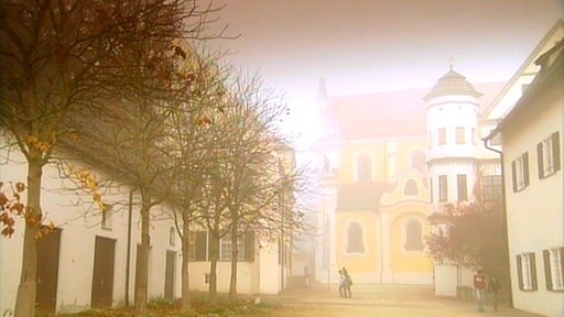 Kloster Ochesenhausen, mehrere Gebäude und in einer Reihe stehende Bäume. Einige Personen laufen über das Gelände.