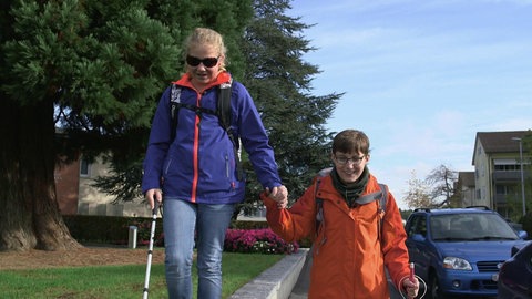 Rebekka und Florian laufen händehaltend mit Blindenstöcken die Straße entlang.