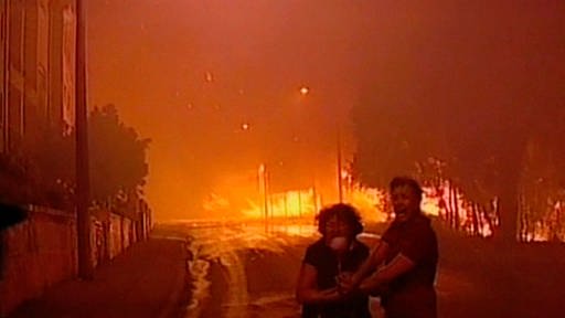 Zwei verzweifelnd aussehende Menschen stehen auf einer Straße in einer brennenden Stadt. Links und rechts sind Häuser zu erkennen, rechts vorne Bäume. Das Bild ist vom Feuer orange erleuchtet. (Foto: SWR/WDR – Print aus der Sendung)