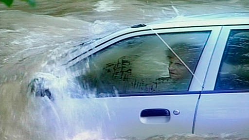 Blick auf die Fahrerseite eines Autos, das gerade von Wassermassen überspült wird: Am Steuer sitzt eine ältere Frau. (Foto: SWR/WDR – Print aus der Sendung)