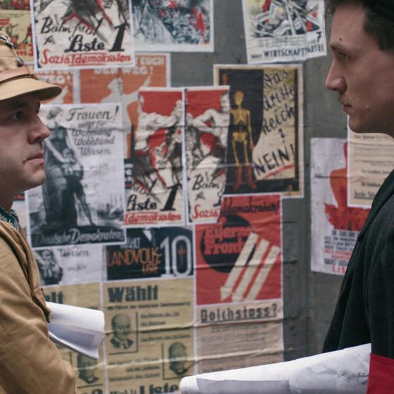 Ein Mann in brauner SA Uniform (links) und ein Mann in Anzug und Hakenkreuzbinde (rechts) vor einer Plakatwand. (Foto: SWR - Screenshot aus der Sendung)