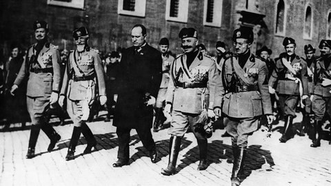 Mussolini beim Marsch auf Rom, er trägt einen schwarzen Mantel und läuft in der ersten Reihe, neben und hinter ihm marschieren viele Männer in Uniform.