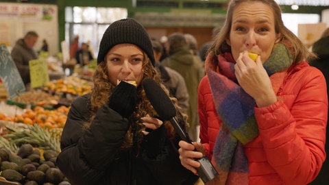 Stefanie und eine Markthändlerin beißen in Zitronenstücke in der Markthalle von Joigny. Welche französischen Begriffe gibt es rund um das Gemüse- und Obstangebot?