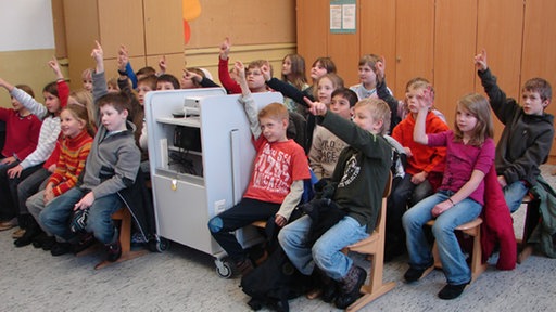 Eine Grundschulklasse sitzt in Stuhlreihen und schaut nach vorn.