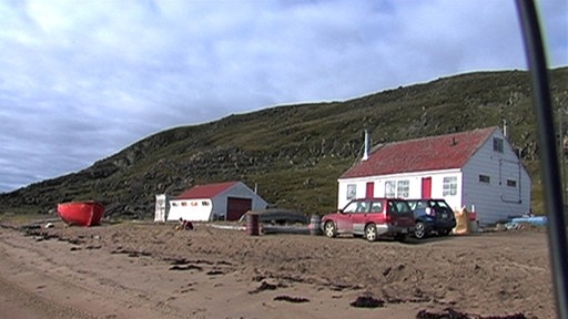 Hudson Bay Company. Mehrere Autos stehen vor zwei Häusern vor einem Hügel.