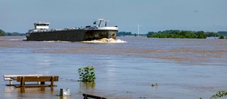 Ein Frachtschiff auf einem Fluss. Der Fluss hat Hochwasser. Im Vordergrund überschwemmte Bänke und eine Person, die aufs Wasser guckt. Welchen Einfluss hat der Mensch auf Flüsse?